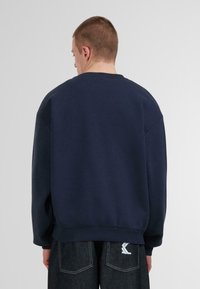 Sudadera azul marino con cuello redondo, corte holgado y puños de canalé. Los detalles incluyen una tela texturizada y un logo visible en la parte posterior.