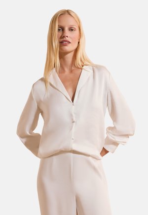 Femme aux longs cheveux blonds porte un chemisier blanc ample boutonné et un pantalon assorti, debout avec les mains dans le dos devant un fond uni.