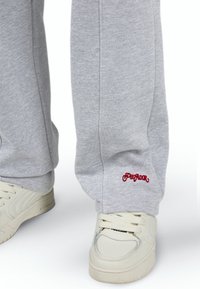 Lys grå sweatpants med rød "PEGADOR" logo over hvide sneakers med snørebånd på en hvid baggrund.