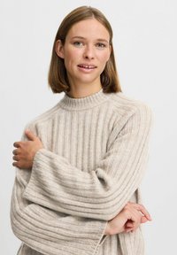 Beige neule paksuilla hihansuilla ja korkealla kauluksella, pehmeä rakenne ja minimalistinen muotoilu. Malli pitää käsiään ristissä.