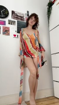 Mlada ženska s kodrastimi lasmi stoji bosonoga, oblečena v pisano prugasto obleko in usklajen plašč, v rokah drži črne visoke pete v notranjosti.