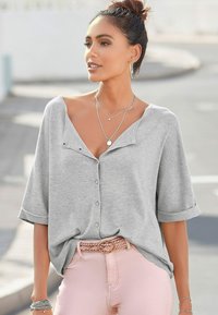 Helles graues kurzärmliges Hemd mit Knopfleiste und V-Ausschnitt, aufgerollten Ärmelenden und weicher Textur; getragen mit rosa Hosen und mehreren Lagen von Halsketten.