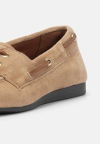 Tan suede slip-on sko med gullfargede snørehull og brune detaljer. Har en avrundet tå og en fleksibel svart gummisåle.