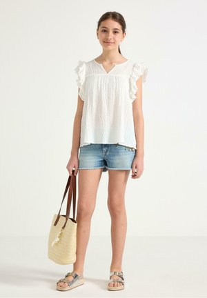 Teenagepige iført hvid flæseærmeløs top, denimshorts, sølv sandaler, holder beige flettet tote-taske med brune håndtag.