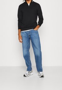 Mann, der einen schwarzen Viertelreißverschluss-Pullover, blue Jeans und weiße sowie silberne Sneakers trägt, steht vor einem schlichten hellen Hintergrund.