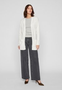 Cardigan largo blanco con bordes acanalados y dos bolsillos delanteros, combinado con una camiseta gris y pantalones de pierna ancha oscuros, junto con tacones.