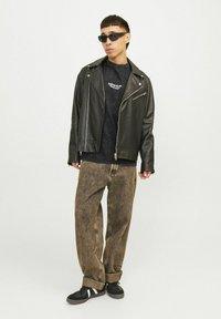 Sort læderbikerjakke med sølvfarvede lynlåse over en mørk t-shirt, parret med lysebrune baggy jeans og sorte sneakers med hvide detaljer.
