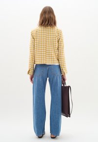 Gul och vit houndstooth-mönstrad jacka, kort design, kombinerad med ljusblå vida jeans. En mörkbrun handväska hålls i ena handen.