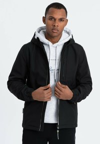 Veste noire en matériau léger avec capuche, fermeture éclair à l'avant et poignets élastiques. Portée par-dessus un sweat à capuche gris avec un design textuel.