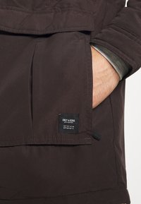 Tmavě hnědá bunda s přední kapsou, na které je černá nášivka. Materiál vypadá hladce s lehkým leskem; design je jednoduchý a funkční.