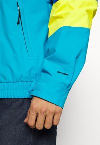 Niebieska wodoodporna kurtka z żółtymi akcentami, elastycznymi mankietami i boczną kieszenią na suwak. Na rękawie widoczne logo "DRYVENT". Gładka faktura.