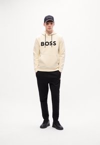 Jonge man in een crème hoodie met BOSS-logo, zwarte joggers, zwarte sneakers en een zwarte BOSS-pet, staande tegen een witte achtergrond.