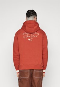 Röd Nike-hoodie med stor "Just Do It"-tryck på baksidan, med dragsko i huvan och ribbade muddar. Mjuk tygkvalitet med en avslappnad passform.