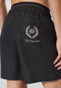 Crne kupaće hlače s elastičnim strukom, s stražnjim džepom ukrašenim bijelim logotipom i lovorovim uzorkom.