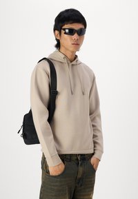 béžová mikina s kapucňou a šnúrkami, čierna crossbody taška, tmavé slnečné okuliare a modré džíny s jemným vertikálnym vzorom.