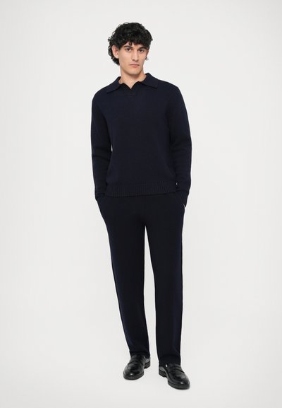 Marinblå tröja med krage, ribbade ärmslut, som matchas med byxor i samma färg. Texturerat tyg. Modellen står med händerna i fickorna. Svarta skor.
