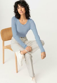 Světle modrý top s dlouhým rukávem, krémové pohodlné kalhoty a bílé sneakers. Model sedící na dřevěné židli, předvádějící casual outfit.