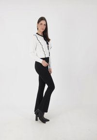 Biała bluzka z czarną falistą lamówką i guzikami, zestawiona z rozkloszowanymi czarnymi spodniami i czarnymi botkami na obcasie, na gładkim tle.