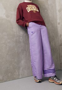 Vinröd sweatshirt med texten "Adidas Originals", kombinerad med lösa ljuslila byxor med tre ränder och en Adidas-logotyp. Svarta och orange sneakers.