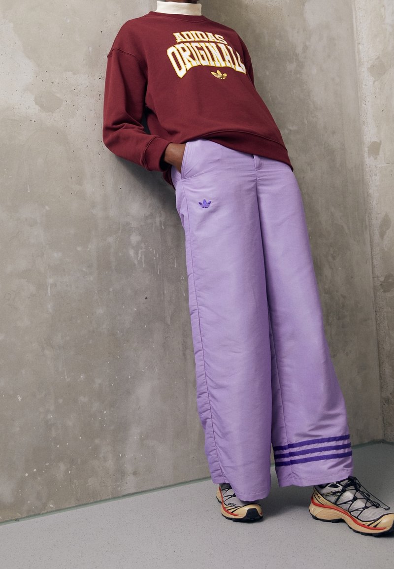 Vinröd sweatshirt med texten "Adidas Originals", kombinerad med lösa ljuslila byxor med tre ränder och en Adidas-logotyp. Svarta och orange sneakers.