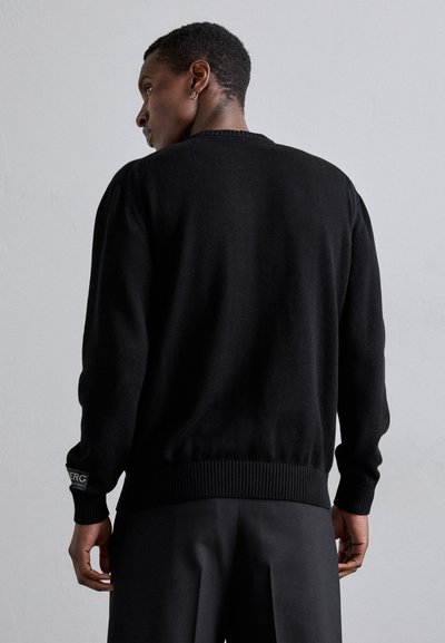 Pull en tricot noir avec un col rond, des bords côtes aux manches et à l'ourlet. Détail logo sur le poignet gauche. Texture lisse, design ajusté.