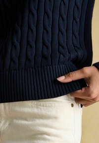 Detailní záběr ruky, která upravuje lem tmavěmodrého svetr s pleteným vzorem přes krémové kalhoty.