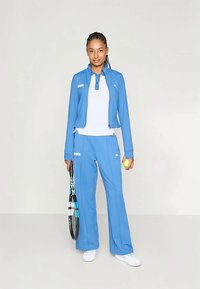 Μπλε αθλητική φόρμα με φερμουάρ και ταιριαστά παντελόνια. Λευκό εσωτερικό μπλουζάκι, ρακέτα τένις και μπάλα στα χέρια. Λείο ύφασμα, άνετη εφαρμογή.