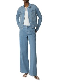 Lichtblauwe spijkerjack en wijde jeans. Het jack heeft twee borstzakken en metalen knopen. Gecombineerd met een aansluitend wit topje en sleehakken.