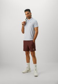 Hele sinine polo särk, millel on peen logo, kombineeritud maroonide lühikesed püksid ja valged tossud. Mudel hoiab käes reketti, demonstreerides spordirõivastust.