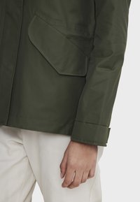Veste vert olive avec une finition texturée, comportant une grande poche à rabat sur le haut à gauche et une manche proprement retroussée, portée sur un pantalon blanc.