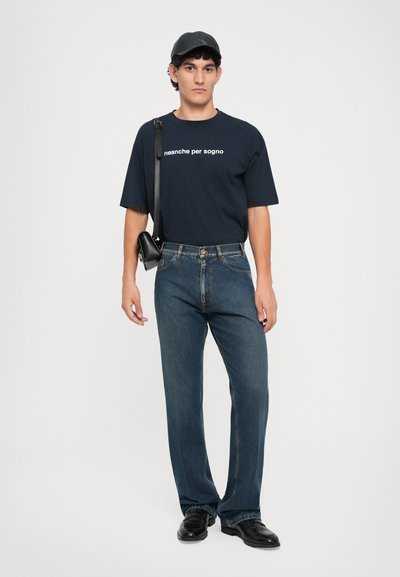 Ung man som bär svart keps, marinblå t-shirt med vit text, blå jeans, svarta skor och liten svart axelväska, stående mot vit bakgrund.
