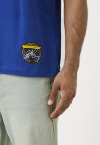 Tricou din bumbac albastru cu un logo rotund care afișează "Steph Curry" în negru și galben în colțul stâng jos. Croiala este casual și relaxată.