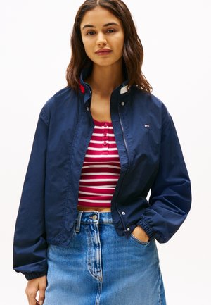 Chaqueta bomber azul marino con cierre frontal y cuello alzado. Combinada con una blusa de rayas rojas y blancas y una falda de mezclilla de cintura alta.