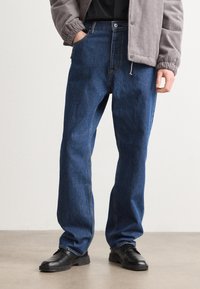 Mørkeblå denim jeans med en afslappet pasform, der har et traditionelt fem-lomme design og synlig syning. Bæres med sorte sko.