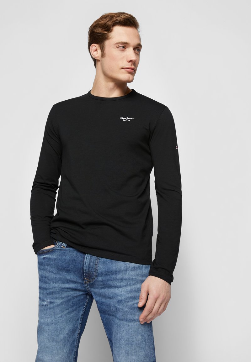 Schwarzes Langarm-T-Shirt aus Baumwolle, mit einem kleinen weißen Logo auf der Brust und taillierter Passform. Getragen mit blauen Jeans.