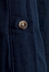 Tissu marine avec un tissage texturé, comportant un bouton en mat marron clair embossé avec "SOLID" et quatre boutonnières.