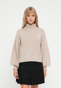 Pulover turtleneck bej deschis, cu o textură moale și pufoasă și mânici bufante, purtat cu o fustă plisată neagră, cu dungi albe.