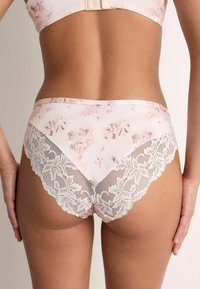 Sieviete valkā gaiši rozā ziedu mežģīņu biksītes ar mežģīnēm un atbilstošu krūšturi, redzama no muguras uz nevienmērīga fona.