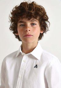 Witte overhemd met opgestaan kraag, voorzien van een klein marineblauw geborduurd ontwerp van doodskop en gekruiste botten op de linkerborst.