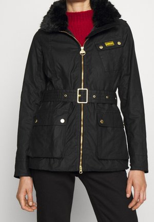 Czarna kurtka z paskiem, złotym suwakiem i guzikami, kieszeniami z klapami oraz kołnierzem z sztucznego futra, noszona na czerwonym swetrze przez osobę z rozluźnionymi rękami.