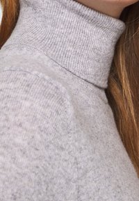 Närbild av en person som bär en ljusgrå rippad polotröja med långt brunt hår synligt på sidan.