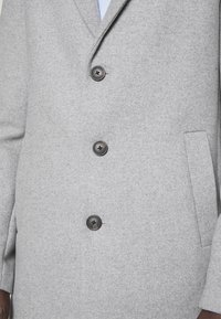 Jasnoszary wełniany płaszcz z trzema czarnymi guzikami zapiętymi do góry i ukośnymi bocznymi kieszeniami, noszony na białej koszuli.