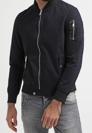 Mężczyzna ubrany w czarną kurtkę typu bomber z suwakiem z przodu i zamkniętą kieszenią na lewym rękawie, w połączeniu z jasnoszarymi dżinsami.