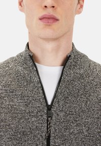 Graue Zip-Jacke mit strukturiertem Strickdesign, schwarzem Reißverschluss, hohem Kragen und weißem Innen-Shirt, das unter der Jacke sichtbar ist.