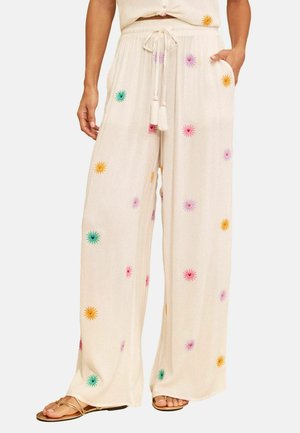Femeie care poartă pantaloni largi crem cu floarea-soarelui multicolor brodată, talie cu șnur, buzunare laterale și sandale aurii.
