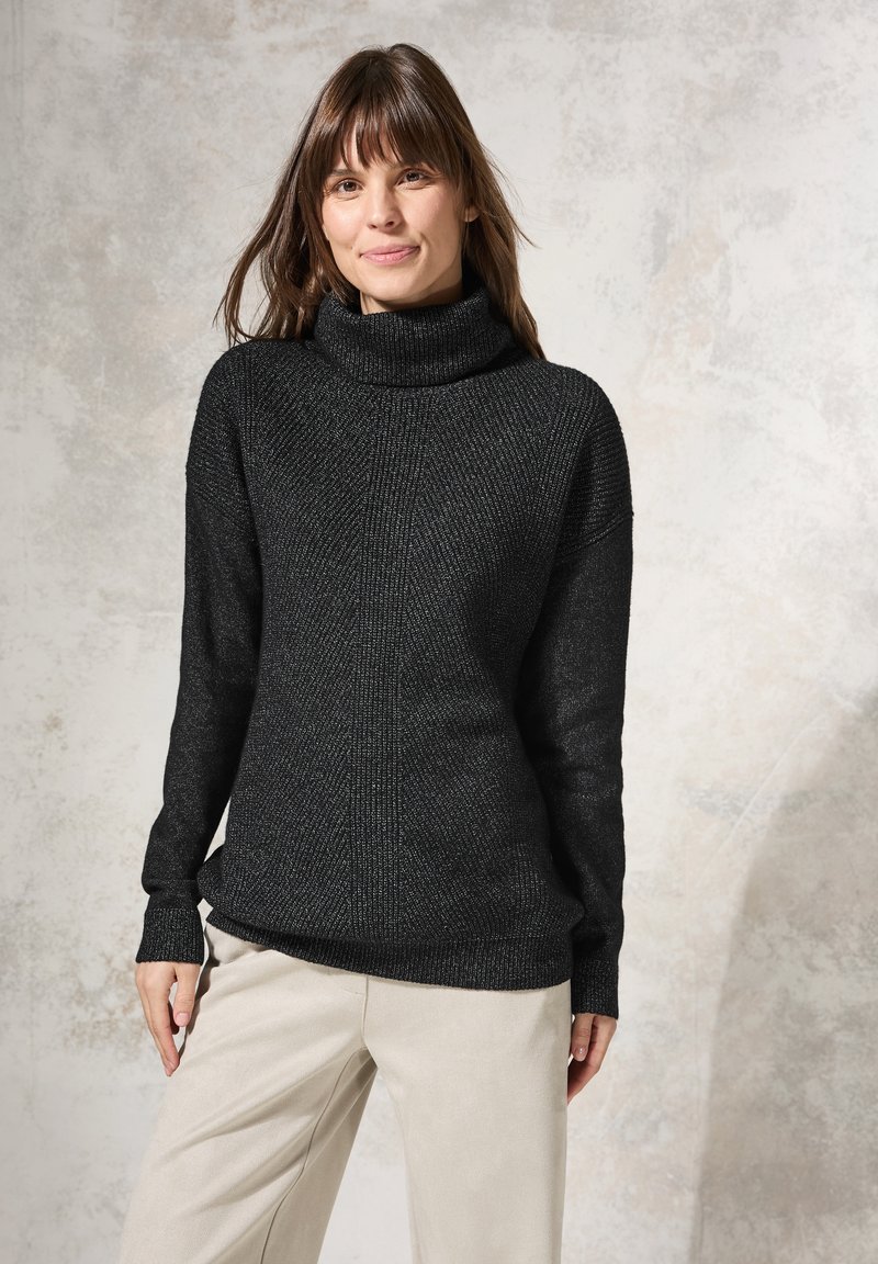 Maglione a collo alto nero con un design a maglia testurizzato, caratterizzato da una vestibilità ampia e polsini a coste. Indossato sopra pantaloni chiari.