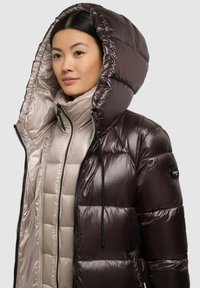 Braune wattierte Jacke mit glänzendem Außenmaterial, großer Kapuze, Frontreißverschluss und beigem Innenfutter. Verfügt über eine gesteppte Textur und ein Logo-Patch.