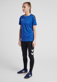 Blå sport t-shirt med kort ärm och vit krage, matchad med svarta leggings med vita chevron-mönster. Blå sportskor fulländar outfiten.