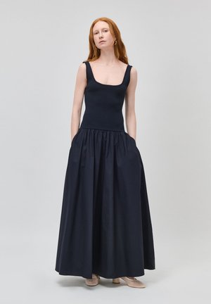 Femme aux longs cheveux roux portant un haut noir sans manches et une longue jupe noire avec poches, debout devant un fond clair uni.