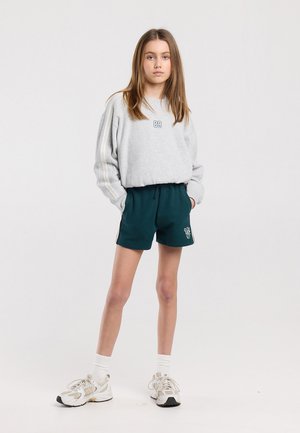 Jeune fille debout les mains dans les poches, portant un sweat-shirt gris et un short de sport vert foncé avec des baskets blanches sur un fond blanc.